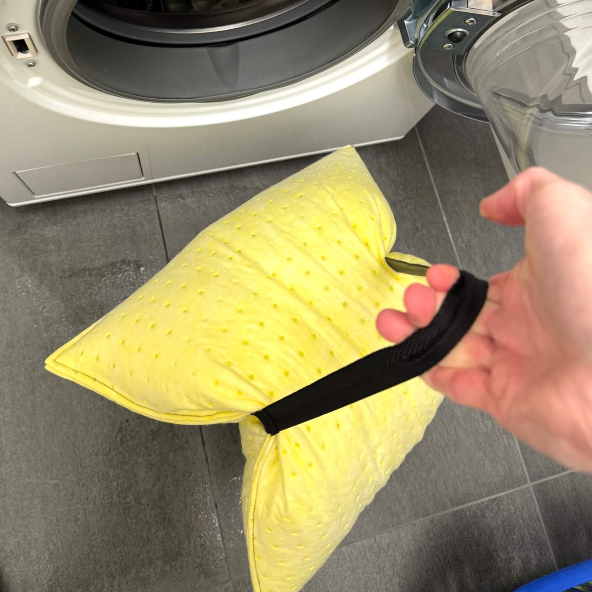 Eine Hand trägt das vollgesogene Kissen mit dem Tragegurt von der Waschmaschine weg